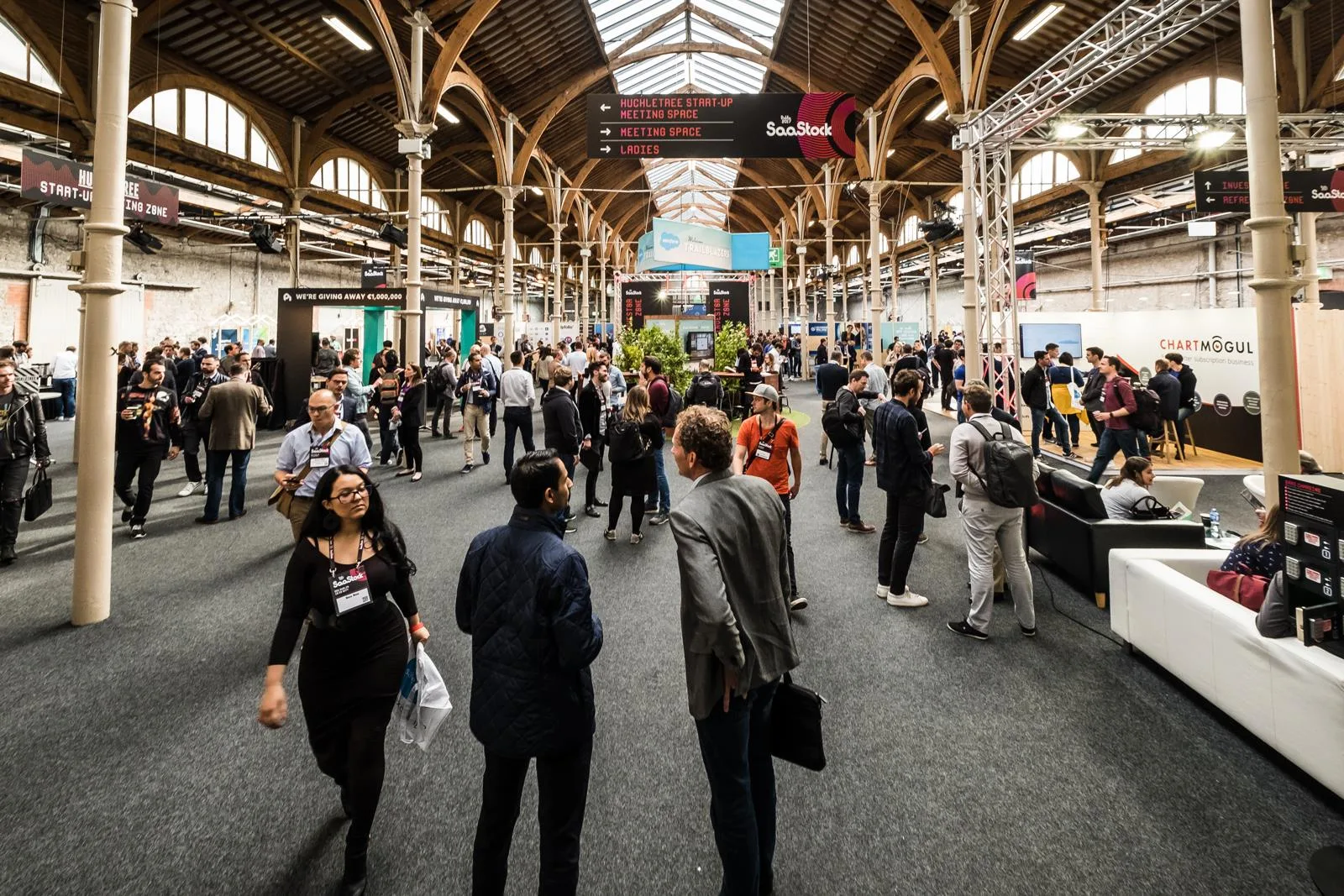 Live at SaaStock Europe 2017 — wide expo-floor view of the RDS Industries Hall with ChartMogul's sponsor stand on the right, the Huckletree Start-Up Zone signs overhead, and attendees moving between booths