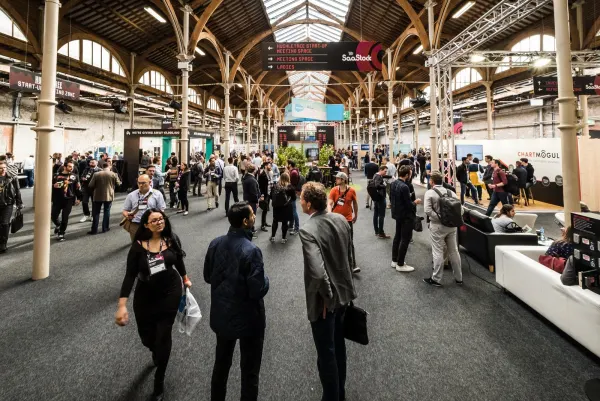Live during SaaStock Europe 2017 — wide expo-floor view of the RDS Industries Hall in Dublin: attendees streaming between booths under the original Victorian brick-and-iron timber roof, with the ChartMogul sponsor stand on the right, the Huckletree Start-Up Meeting Space signs hanging overhead and a SaaStock wayfinding marker pointing to the Meeting Space and Ladies