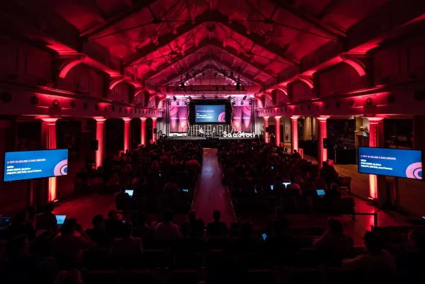 Live during SaaStock Europe 2017 — rear-of-house view of the SaaStock mainstage at RDS Industries Hall, Dublin: red uplighting along the original Victorian timber-vaulted roof, packed seated audience down the centre aisle, and twin side screens showing the slide '5 Tips to Selling a SaaS Unicorn — Mike Davis, Qualtrics; Philippe Botteri, Accel Partners'