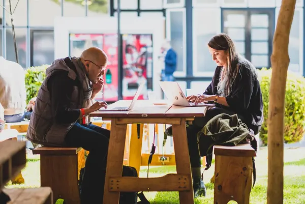 Live during SaaStock Europe 2017 — two attendees working from laptops at a wooden picnic table in the outdoor courtyard between RDS halls, Dublin, with greenery, daylight and other attendees visible in the background
