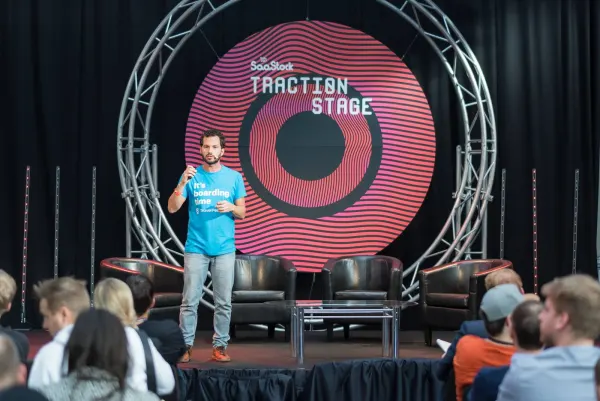 Live during SaaStock Europe 2017 — speaker in a teal TravelPerk 'It's boarding time' tee on the SaaStock Traction Stage in Dublin, mic in hand, in front of the red SaaStock circular Traction Stage backdrop with three empty leather armchairs behind him and a low coffee table on stage