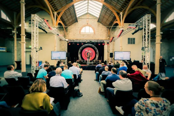 Live during SaaStock Europe 2017 — Traction Stage in a side hall at RDS Simmonscourt, Dublin: a speaker centre-stage in front of the SaaStock-branded red circular backdrop, two flanking IMAG side screens, lighting truss arch overhead and a seated audience under the original Victorian timber roof