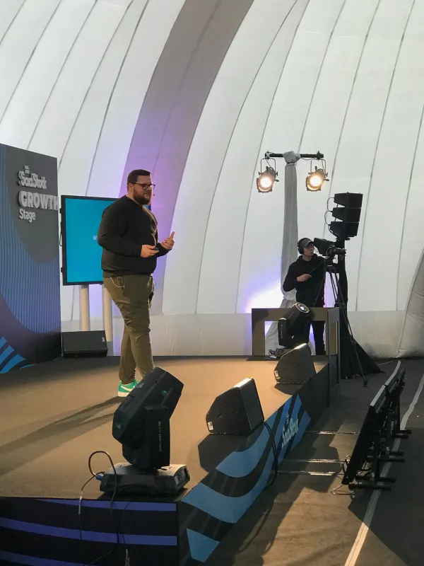 Behind the scenes at SaaStock Europe 2018 — portrait of the Growth Stage with a cameraman filming from the SaaStock-wave-branded stage edge, lighting trusses overhead, and the 'SaaStock Growth Stage' backdrop signboard visible at left