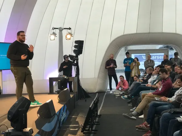 Live during SaaStock Europe 2018 — Growth Stage speaker mid-talk in front of a 'we fail at scale' side-screen slide, with a camera operator filming on a tripod at stage right and attendees in a 'Larsen Contractors' branded tee visible past the dome doorway