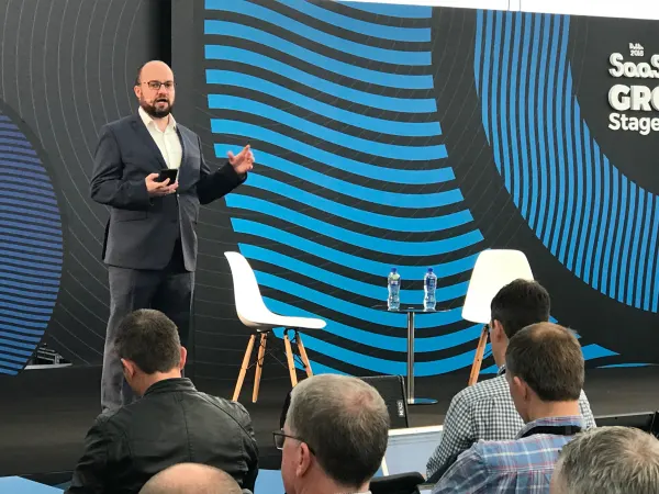 Live during SaaStock Europe 2018 — Thomas Smale mid-gesture on the SaaStock Growth Stage in front of the blue wave 'SaaStock Growth Stage' curved LED backdrop, with two empty Eames chairs behind him