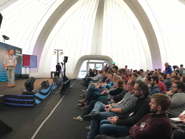 Live during SaaStock Europe 2018 — Growth Stage audience-side angle: speaker at left, bullet-point slide on the side screen, and ~80 attendees in seated rows under the dome