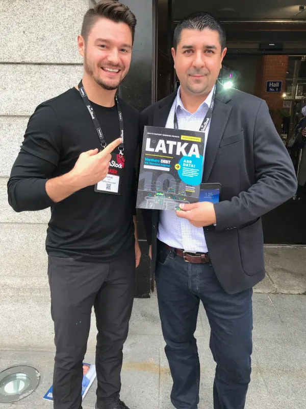 Live during SaaStock Europe 2018 — Nathan Latka in black with a SaaStock lanyard pointing at the LATKA Magazine being held by an attendee in a grey suit outside Hall No. 1 at RDS Simmonscourt, Dublin