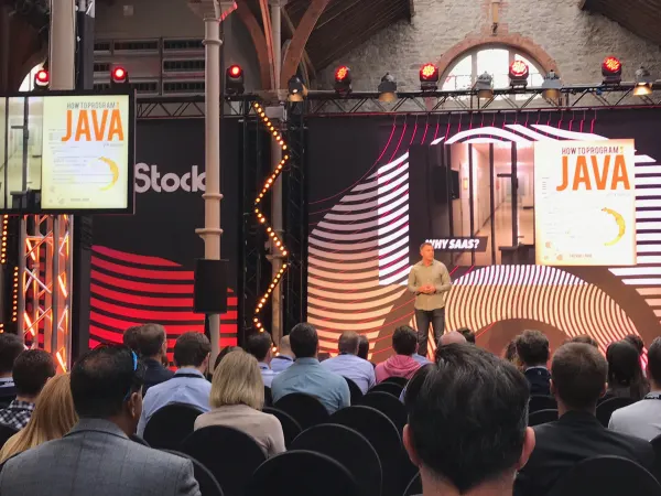 Live during SaaStock Europe 2018 — closer view of the Pitch Stage with the 'WHY SAAS?' wave-pattern LED backdrop and the 'How to program Java' book-cover gag repeated on the side screens