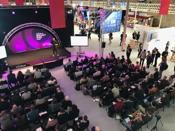 Live during SaaStock Europe 2018 — wider balcony view of the Rocket Fuel Stage showing the full Industries Hall context, attendees streaming past at the back of the room and floor signage above the booths