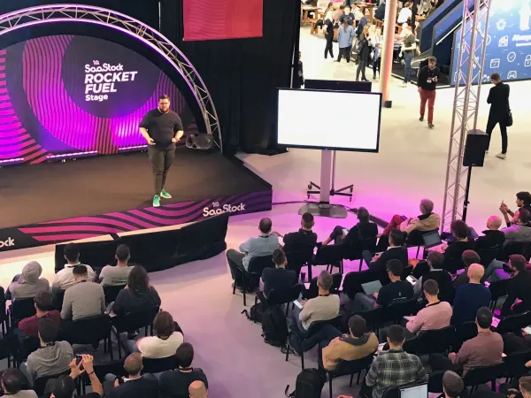 Live during SaaStock Europe 2018 — side angle of the Rocket Fuel Stage with the speaker mid-walk, a blank secondary IMAG screen on its own tripod, and 'SaaStock' branding along the stage step