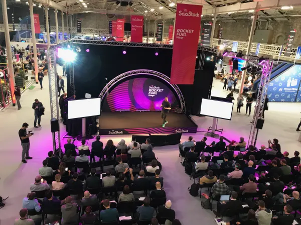 Live during SaaStock Europe 2018 — wide overhead frame from the RDS Industries Hall balcony showing the Rocket Fuel Stage centred, the brick walls and original timber roof in full view, and a red SaaStock banner hanging overhead