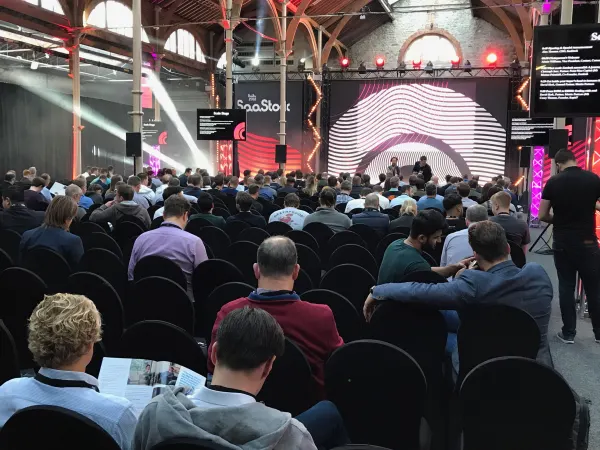 Live during SaaStock Europe 2018 — Scale Stage in the RDS Industries Hall, Dublin: pink wave LED backdrop, two figures on stage, and a packed audience visible in the seated rows under the iconic brick-and-iron timber roof