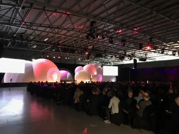 Live during SaaStock Europe 2019 — Mainstage at RDS Simmonscourt seen from the rear of the auditorium, with the full inflatable-balloon stage set lit pink and purple under truss lighting and a large seated audience filling the hall, Dublin