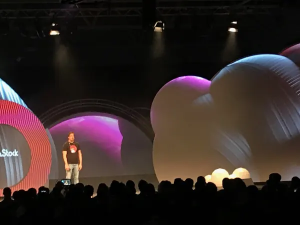 Live during SaaStock Europe 2019 — Mainstage speaker in a black SaaStock-branded tee standing in front of the pink-lit ring screen and inflatable balloons, audience silhouettes in the foreground, Dublin