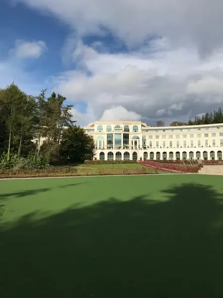Powerscourt Hotel Resort & Spa exterior — curved cream Palladian facade framed by autumn trees and a wide green lawn under a blue Irish sky