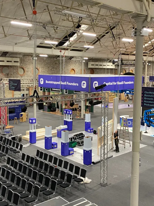 Before doors at SaaStock Europe 2022 — Bootstrap Stage area mid-build with empty black chairs, hanging 'Bootstrapped SaaS Founders' / 'Capital for SaaS Founders' banners, and Founderpath plinths being placed