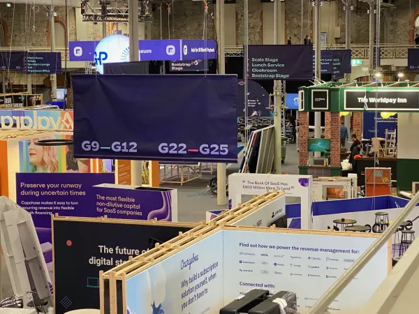 Before doors at SaaStock Europe 2022 — expo aisle with Capchase 'Preserve your runway during uncertain times' panel and Chargebee 'Why build a subscription solution yourself' wall, overhead booth-zone signage 'G9–G12' and 'G22–G25'