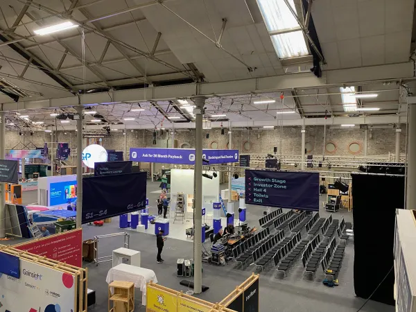 Before doors at SaaStock Europe 2022 — balcony overview of the SaaStock Dublin expo hall mid-build with hanging 'Ask for 36 Month Payback', 'Bootstrapped SaaS Founders', 'Growth Stage / Investor Zone / Hall 4 / Toilets / Exit' wayfinding banners