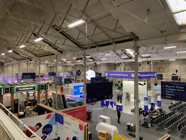 Before doors at SaaStock Europe 2022 — balcony overview of the expo with the 'Worldpay Inn' lounge structure on the left and the Gainsight booth at front-left, build crew still working the floor