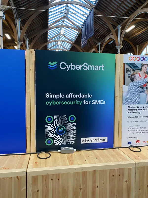 Before doors at SaaStock Europe 2022 — CyberSmart 'Simple affordable cybersecurity for SMEs' booth and the neighbouring Abodoo skills-matching booth on a wooden plinth under the venue's vaulted glass roof