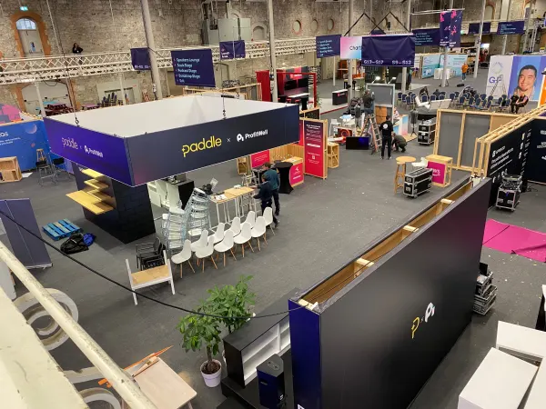 Before doors at SaaStock Europe 2022 — overhead view of the Paddle x ProfitWell sponsor booth at SaaStock Dublin mid-build, white meeting chairs being placed, navy 'Paddle x ProfitWell' branded canopy