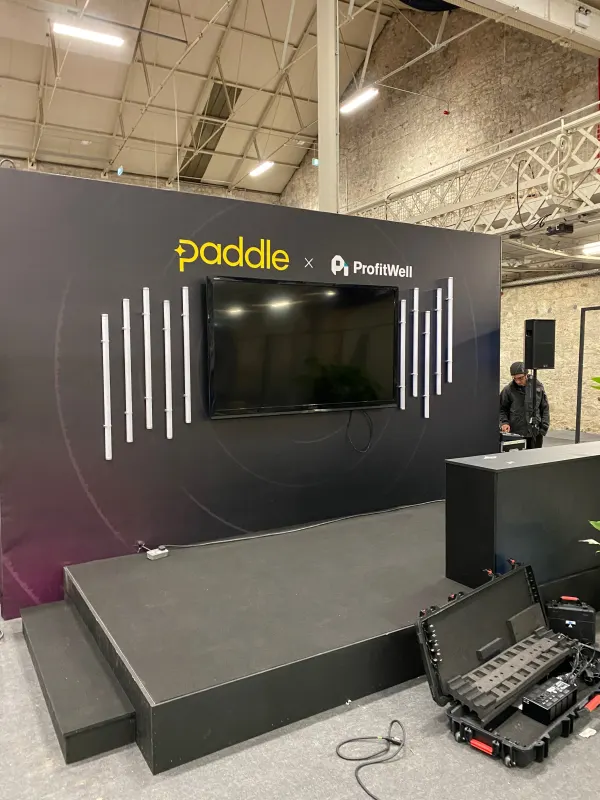 Before doors at SaaStock Europe 2022 — the Paddle x ProfitWell sponsor booth wall mid-build, mounted TV centre, vertical white LED bars on a dark gradient backdrop, road case open in foreground