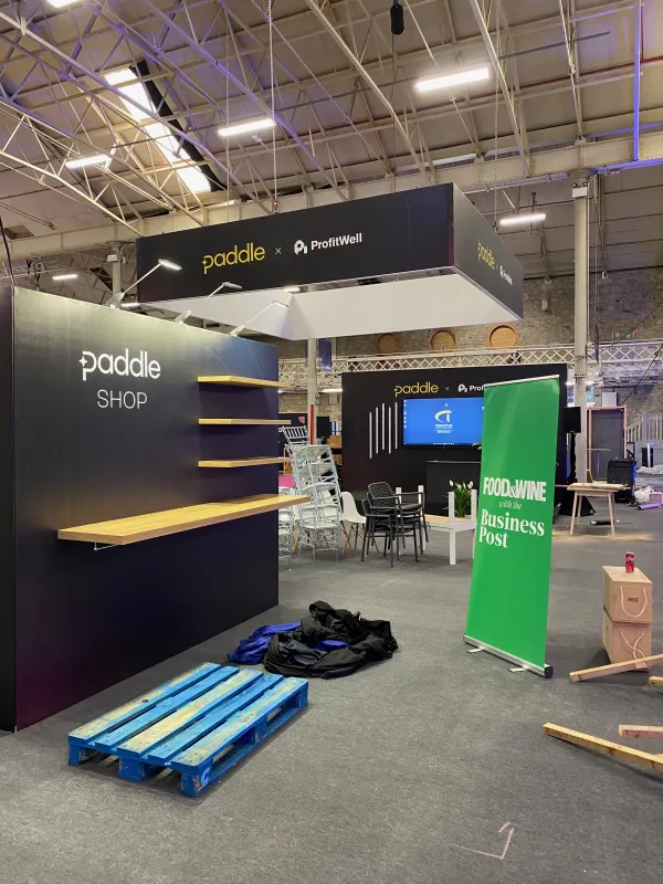 Before doors at SaaStock Europe 2022 — the Paddle Shop swag booth at SaaStock Dublin still being built, empty wooden display shelves, a Food&Wine with the Business Post roll-up next to it