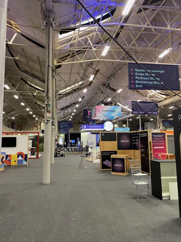 Before doors at SaaStock Europe 2022 — a quiet side aisle in the SaaStock Dublin expo with ChartMogul and Swan booths visible, overhead 'Speakers Lounge / Scale Stage / Podcast Stage / Bootstrap Stage / Exit' signage, no attendees yet