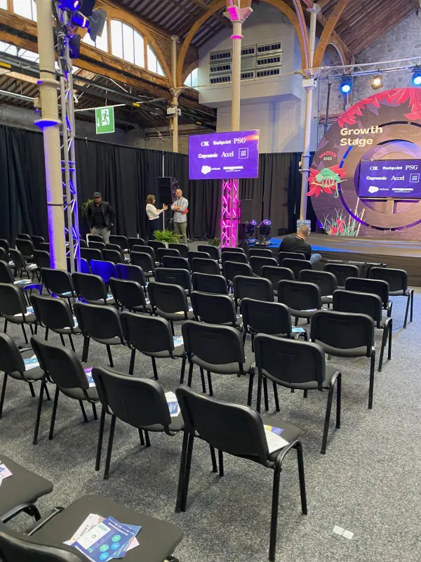 Before doors at SaaStock Europe 2022 — empty Growth Stage at the Dublin venue with the foliage / cactus LED set fully lit, microphones on stand, chairs in rows under the timber-arched roof