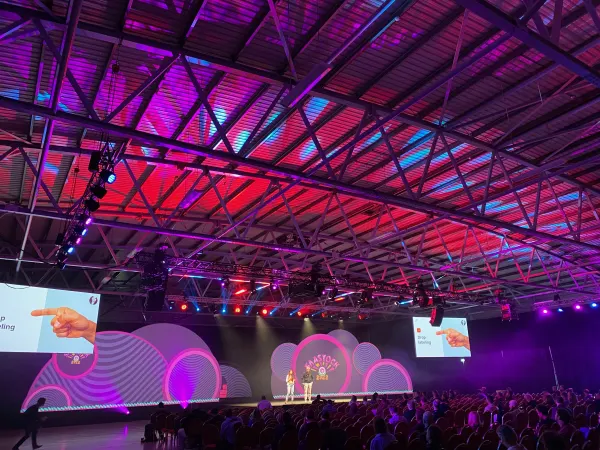 Live during SaaStock Europe 2022 — alternate angle of the SaaStock Dublin Mainstage under the red-and-pink ceiling wash, packed audience watching the presenters in front of the cloud-shaped LED backdrop and pink ring elements