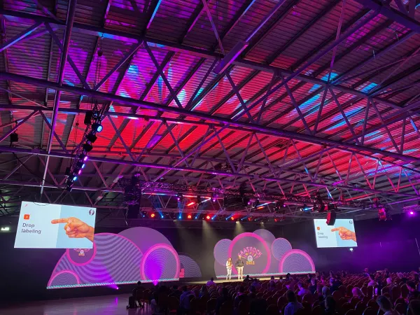 Live during SaaStock Europe 2022 — the SaaStock Dublin Mainstage at full capacity under the venue's red-and-pink lit ceiling, two presenters centre-stage in front of the cloud-shaped SaaStock 2022 LED set, IMAG side screens showing the 'Drop labeling' product slide