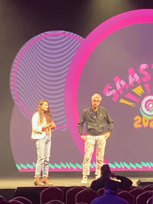 Live during SaaStock Europe 2022 — two speakers standing front-of-stage on the SaaStock Dublin Mainstage in front of the pink cloud-shaped LED set with the green SaaStock 2022 logo visible