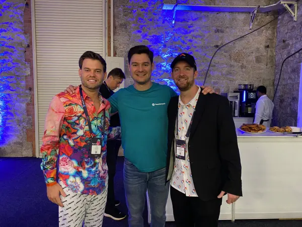 Live during SaaStock Europe 2022 NightStock party — three attendees posing under blue-uplit stone walls, coffee bar visible at right, SaaStock 2022 attendee lanyards on each person