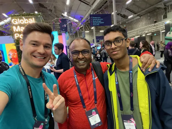 Live during SaaStock Europe 2022 — selfie of three attendees in the expo / networking area with the 'Global' map wall and overhead directional signs (Cloakroom, Lunch Service, Scale Stage, Meeting Tables, First Aid) in the background
