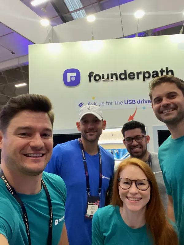 Live during SaaStock Europe 2022 — five members of the Founderpath team in turquoise tees taking a selfie in front of the purple Founderpath sponsor wall with the 'Ask us for the USB drive' lead-capture call-out