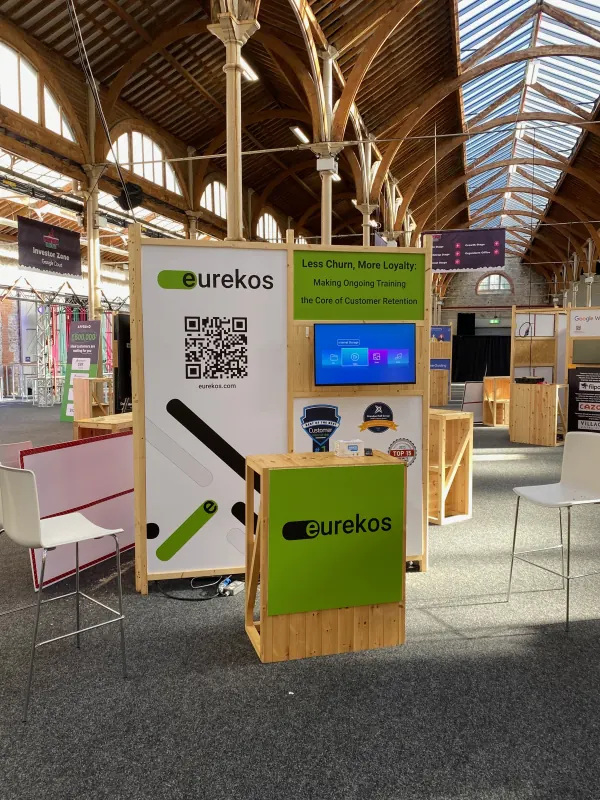 Before doors at SaaStock Europe 2023 — Eurekos LMS sponsor booth with QR code lead-capture and 'Less Churn, More Loyalty' talk teaser under the RDS Simmonscourt timber roof, Dublin