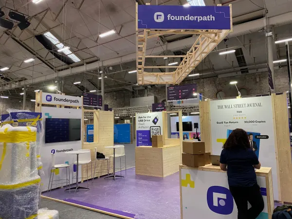 Before doors at SaaStock Europe 2023 — Founderpath sponsor booth being set up in the expo hall, Dublin: purple branding with stools, monitors, and a Wall Street Journal display