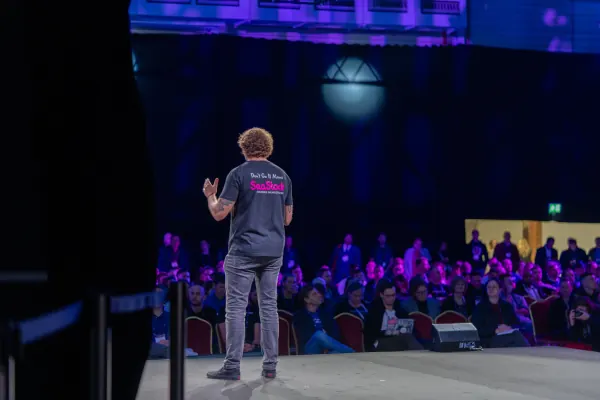 Live during SaaStock Europe 2023 — Mainstage host in a 'SaaStock' branded tee shot from behind on stage, audience visible in the seats at RDS Simmonscourt