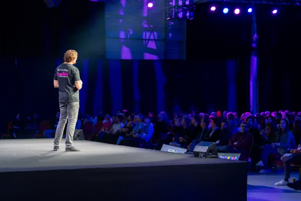 Live during SaaStock Europe 2023 — Mainstage host shot from behind in a SaaStock-branded tee, addressing a full Mainstage audience under blue lighting at RDS Simmonscourt, Dublin