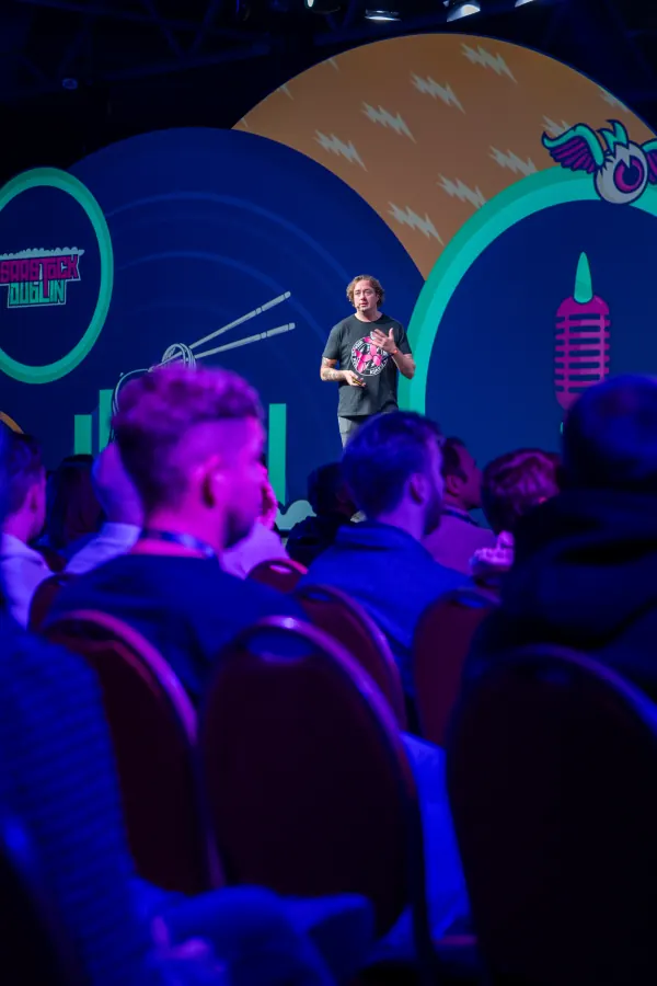 Live during SaaStock Europe 2023 — Mainstage keynote with a speaker in a tee mid-talk, audience silhouetted in blue stage lighting in front of the SaaStock Dublin record-player LED set