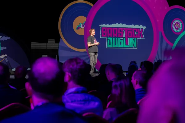 Live during SaaStock Europe 2023 — Mainstage keynote speaker centered against the SaaStock Dublin neon-typography LED backdrop, audience silhouettes in the foreground