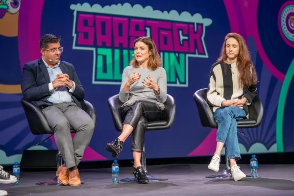 Live during SaaStock Europe 2023 — Mainstage panel of three (two women, one man) seated on chairs with the woman in the middle speaking and gesturing, SaaStock Dublin LED backdrop behind them