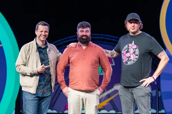 Live during SaaStock Europe 2023 — three Mainstage hosts posing together on the SaaStock Dublin set: one in a tan jacket pointing, one in an orange shirt center, one in a Banksy-balloon-dog black tee with a cap on the right
