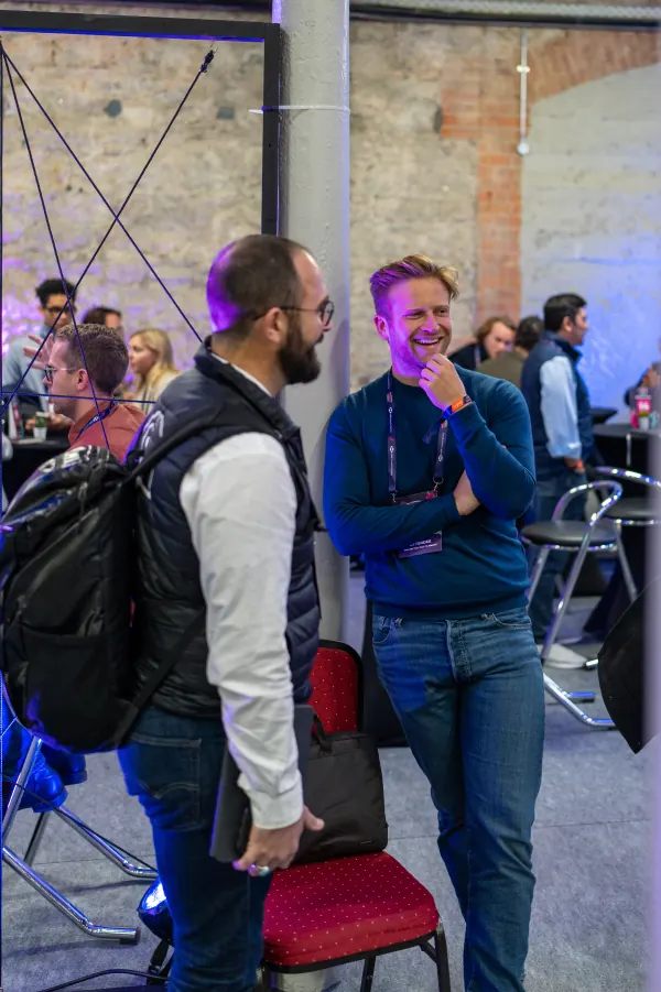 Live during SaaStock Europe 2023 — two founders mid-conversation in the RDS Simmonscourt networking area, one laughing in a teal sweater, the other in a puffer vest with a backpack