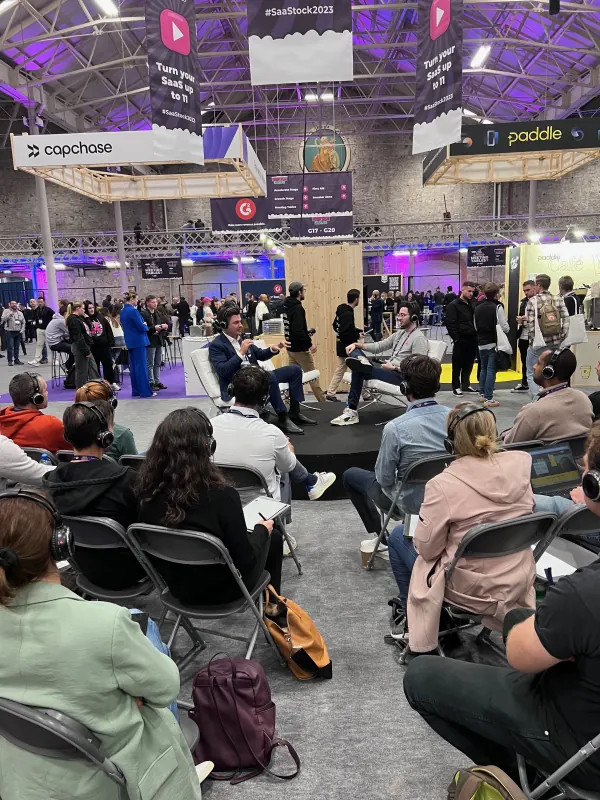 Live during SaaStock Europe 2023 — Podcast Stage panel with three panelists on stools and a seated silent-disco audience on headphones, under Capchase, Paddle and G2 hanging signs