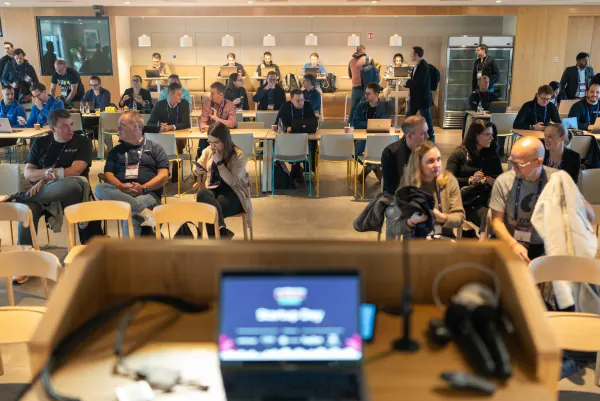 Live at SaaStock Europe 2023 Startup Day, Zendesk Dublin — wide audience shot from the podium POV showing ~25 attendees seated at long tables, with the speaker's MacBook in the foreground running the 'Startup Day' deck