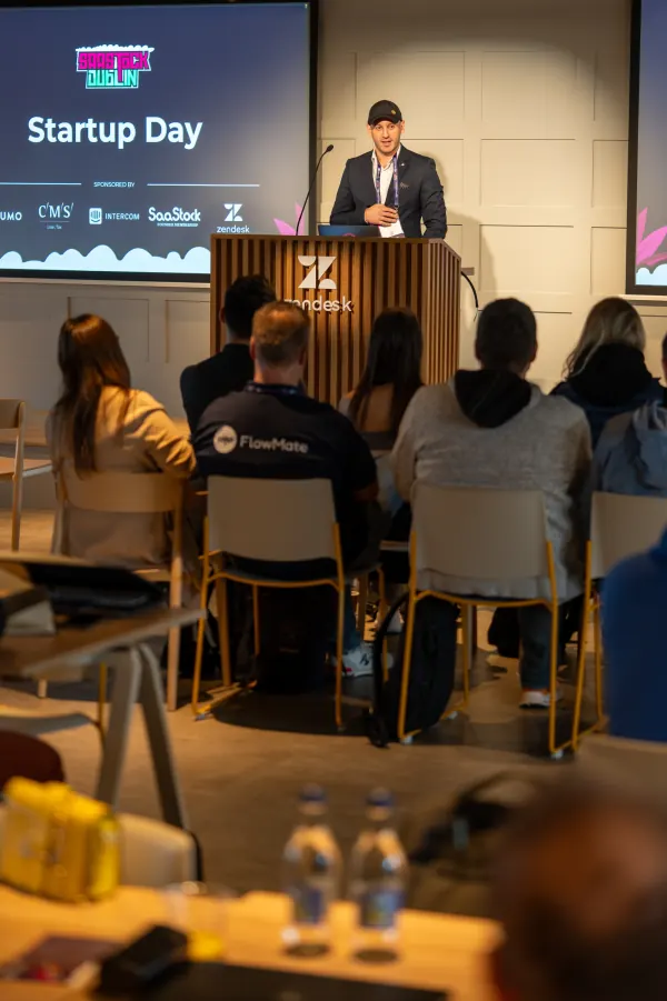 Live at SaaStock Europe 2023 Startup Day, Zendesk Dublin — host on stage shot from inside the audience, with a FlowMate-branded attendee in the foreground and the 'Startup Day' sponsor slide behind