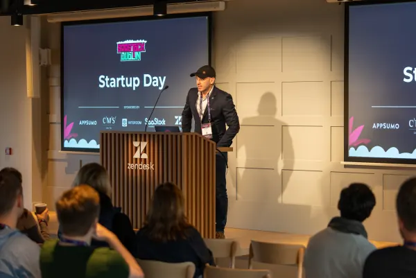 Live at SaaStock Europe 2023 Startup Day, Zendesk Dublin — host in a cap and navy blazer behind the Zendesk podium delivering the opening talk in front of a seated audience