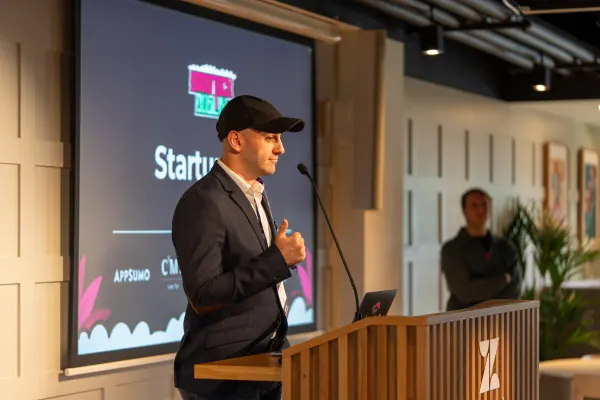 Live at SaaStock Europe 2023 Startup Day, Zendesk Dublin — speaker in a cap and navy blazer mid-talk, side profile, with AppSumo and CMS Hub sponsor logos visible on the slide behind