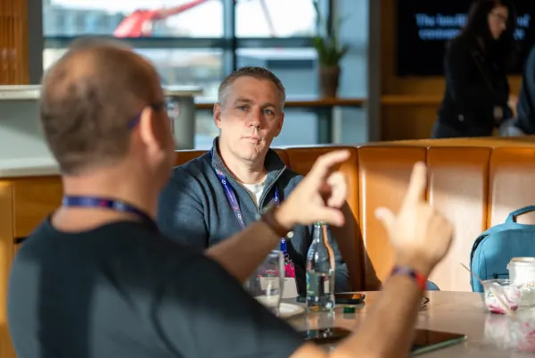 Live at SaaStock Europe 2023 Startup Day, Zendesk Dublin — two attendees mid-conversation across a table, the one closer to camera gesturing with both hands, against a window with a red Dublin crane visible outside